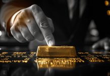Hand tippt auf Goldbarren mit Börsenkursen im Hintergrund